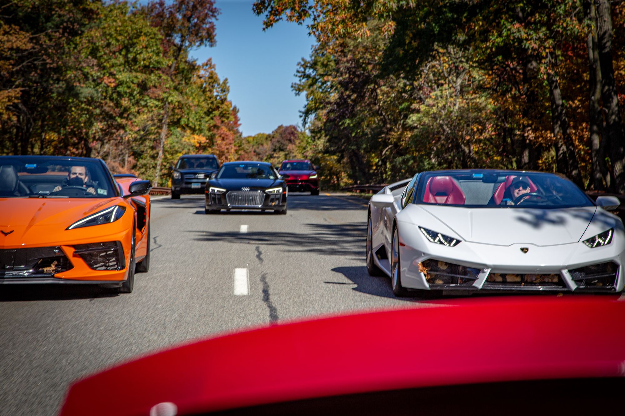 Dream Car Tour convoy on a two-lane road in fall color — an orange Corvette convertible and a white Lamborghini Huracán Spyder lead, an Audi R8 behind