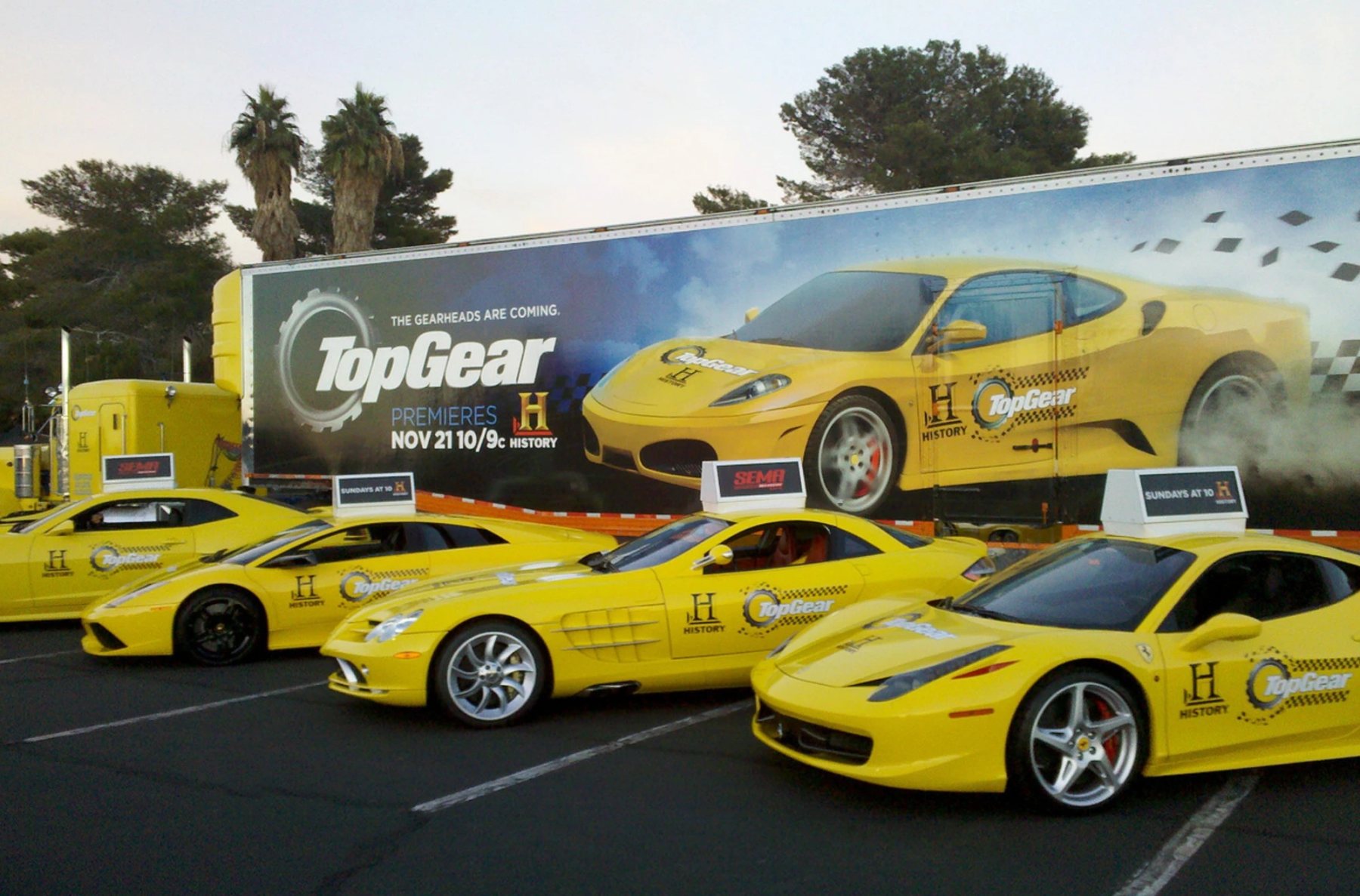 A row of exotic cars wrapped in yellow for a Top Gear premiere promotion, parked in front of the show's trailer